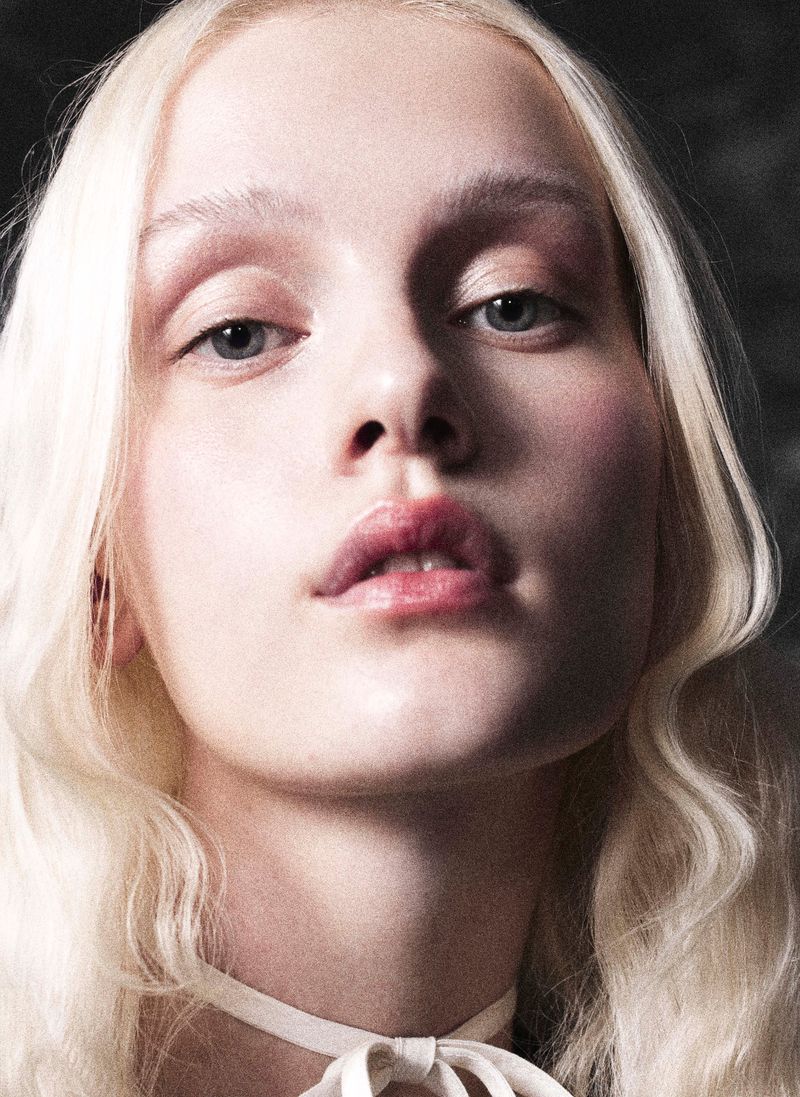 Close-up portrait of a fair-skinned young woman with blonde hair, light eyebrows, and a ribbon choker against a dark background.