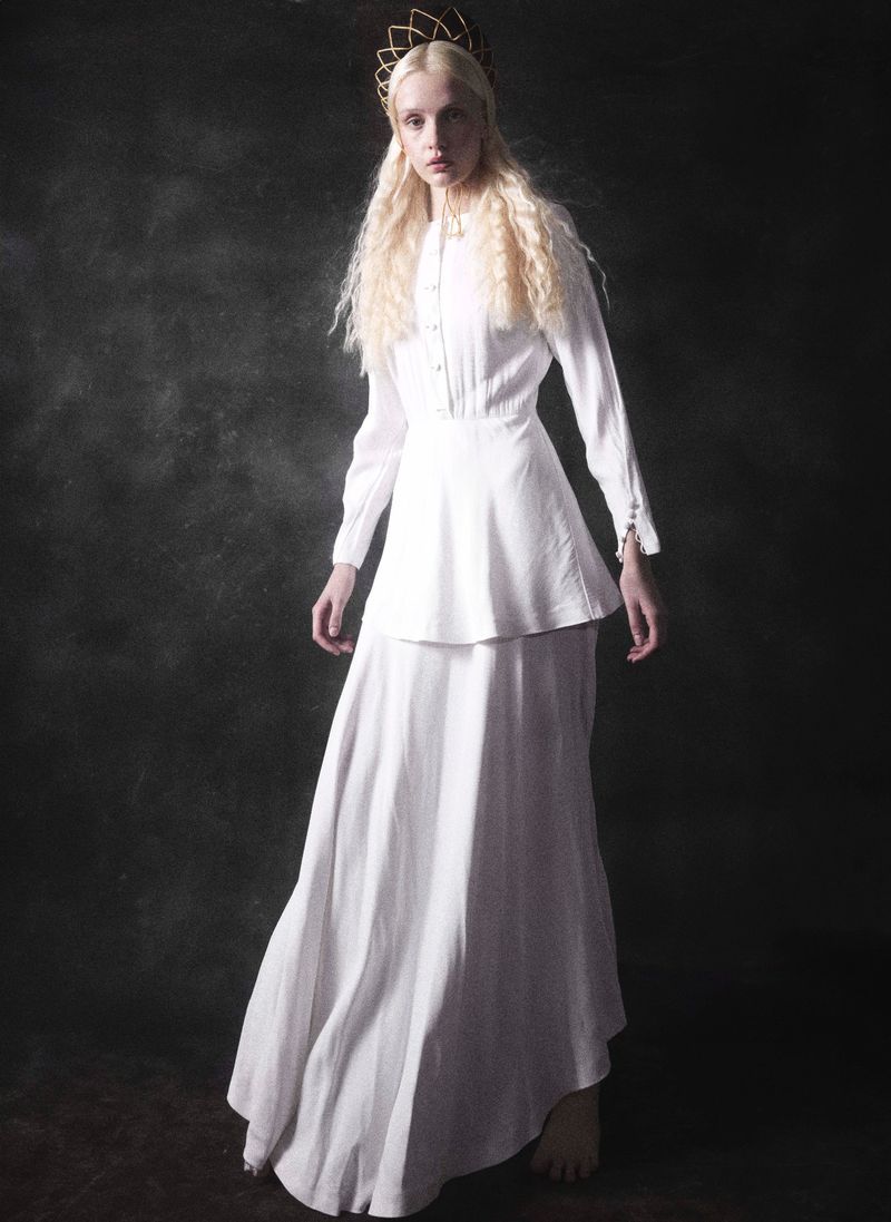 Portrait of an androgynous person wearing a long white dress and gold crown, posing against a dark grey studio background.