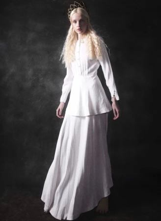 Portrait of an androgynous person wearing a long white dress and gold crown, posing against a dark grey studio background.
