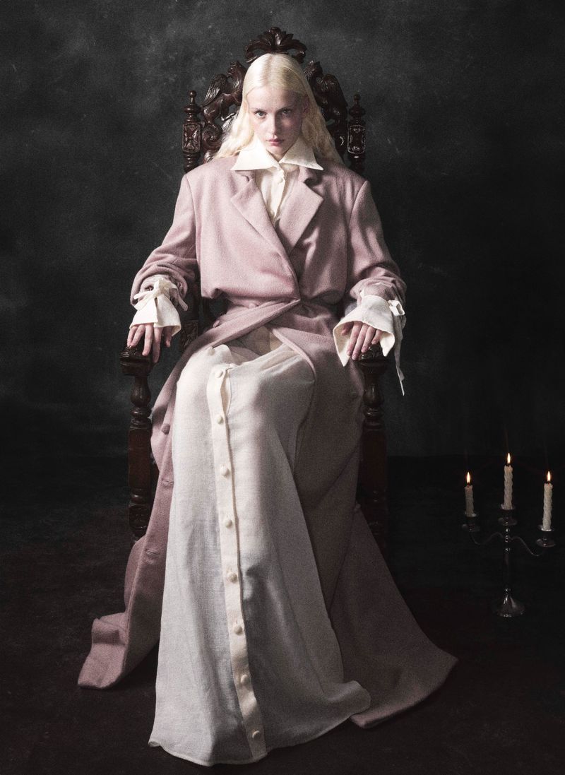 A fair-skinned woman with platinum blonde hair sitting on an ornate wooden throne wearing a light pink coat and white dress.