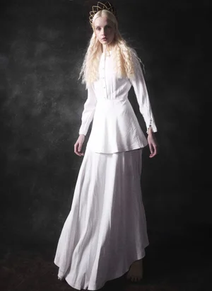 Blonde woman wearing a long white dress and a sculptural crown headpiece posing against a dark background.