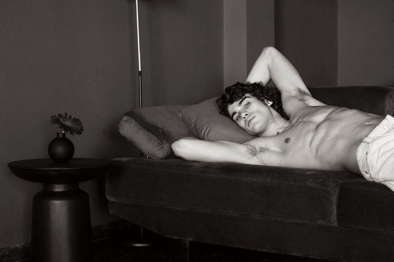 Black and white portrait of a bare-chested adult male lying on a couch in an indoor setting with a modern lamp and flower on a side table.
