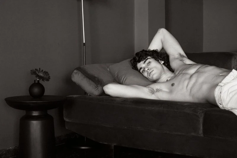 Black and white portrait of a bare-chested adult male lying on a couch in an indoor setting with a modern lamp and flower on a side table.