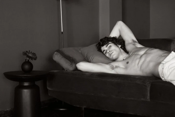 Black and white portrait of a bare-chested adult male lying on a couch in an indoor setting with a modern lamp and flower on a side table.