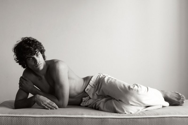 Shirtless man with curly hair wearing light jeans lying down on a mattress in a black and white editorial shoot.