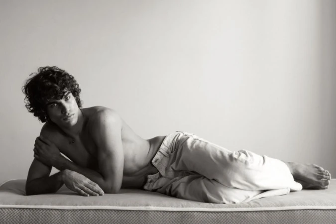 Shirtless man with curly hair wearing light jeans lying down on a mattress in a black and white editorial shoot.
