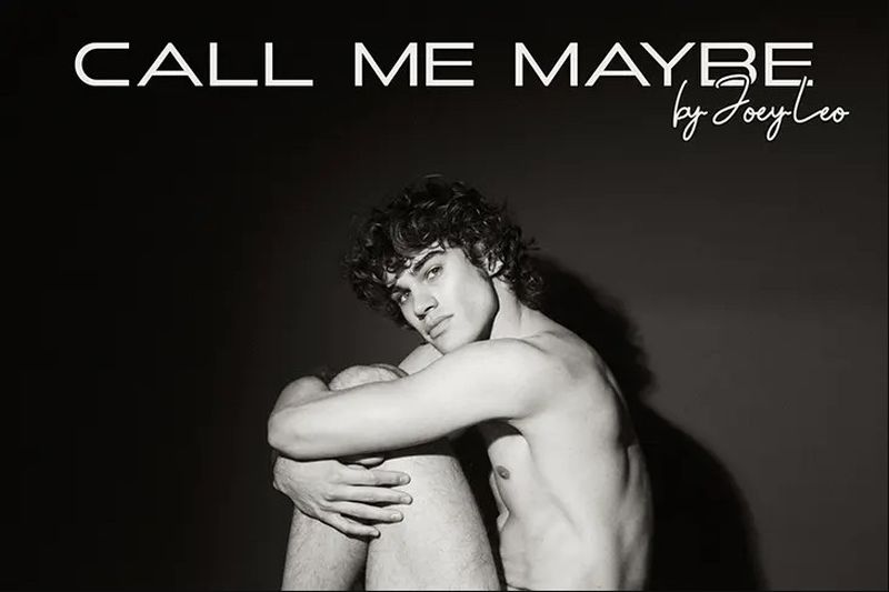 Black and white studio portrait of a shirtless young man with curly hair hugging his knees in a seated pose against a dark backdrop.