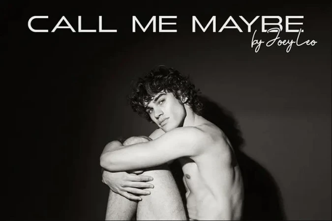 Black and white studio portrait of a shirtless young man with curly hair hugging his knees in a seated pose against a dark backdrop.