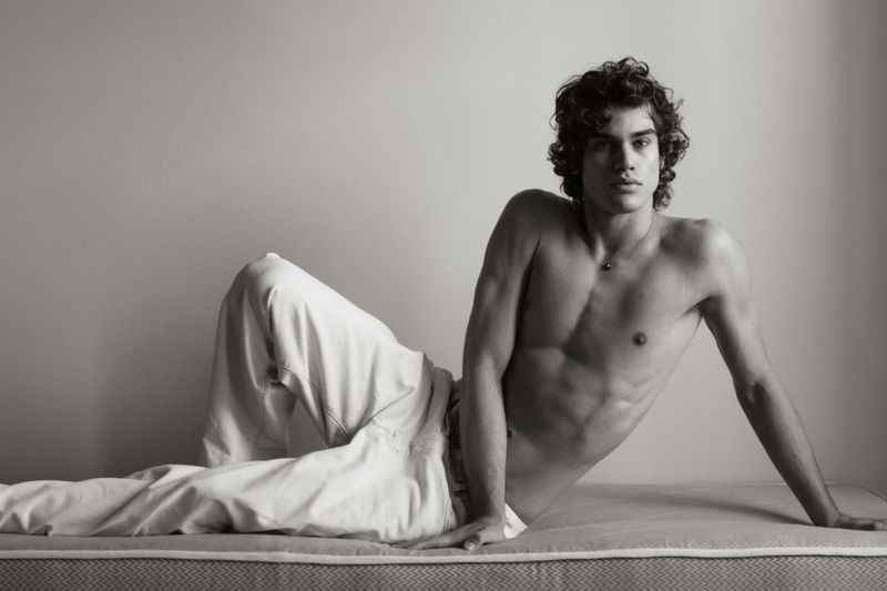 Shirtless young man with dark curly hair reclining on a mattress in white trousers against a plain studio wall.