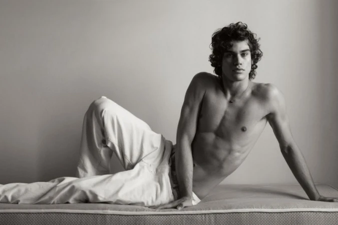 Shirtless young man with dark curly hair reclining on a mattress in white trousers against a plain studio wall.