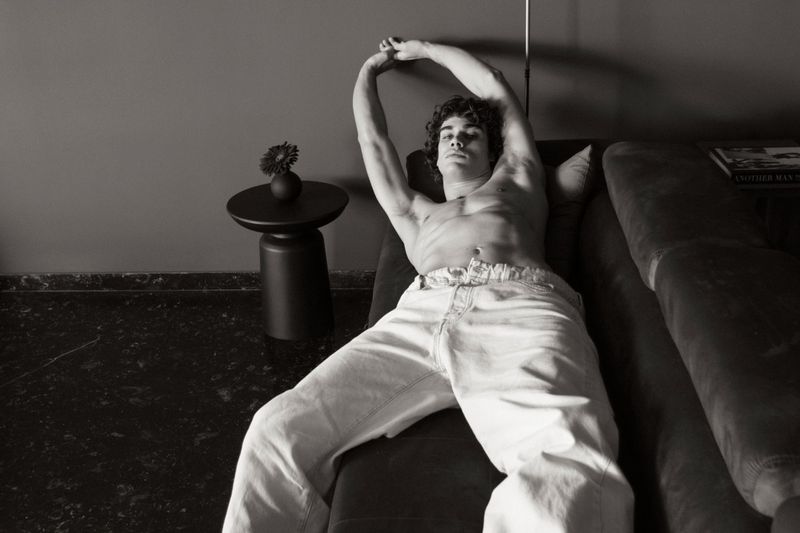 Monochrome image of a shirtless man lying on a couch with arms raised, next to a black side table with a flower in a vase.