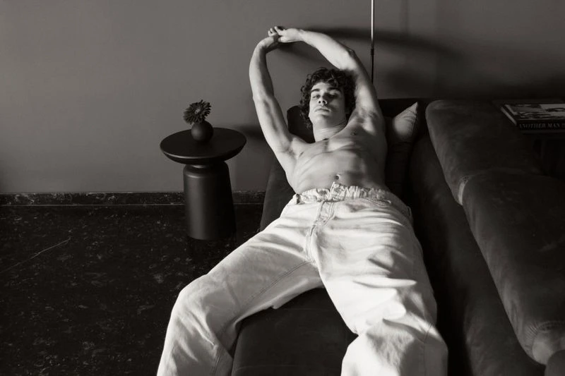 Monochrome image of a shirtless man lying on a couch with arms raised, next to a black side table with a flower in a vase.