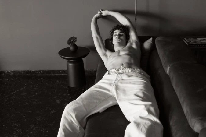 Monochrome image of a shirtless man lying on a couch with arms raised, next to a black side table with a flower in a vase.
