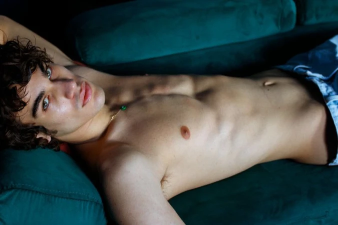 Shirtless young man with curly brown hair poses while lying on a plush teal couch. Gold necklace with green gems is visible.