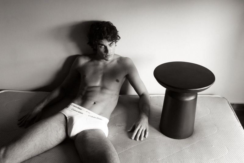 Shirtless man in Calvin Klein underwear laying on a mattress next to a black side table in a black and white studio shot.
