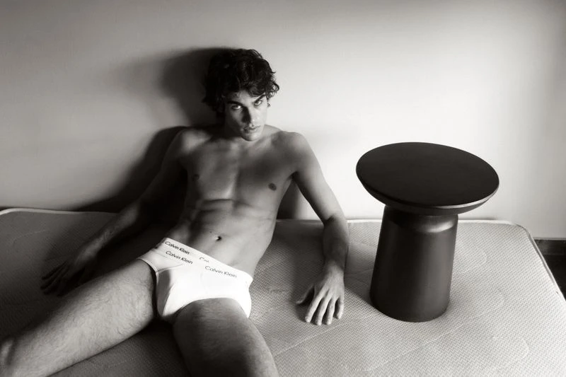 Shirtless man in Calvin Klein underwear laying on a mattress next to a black side table in a black and white studio shot.