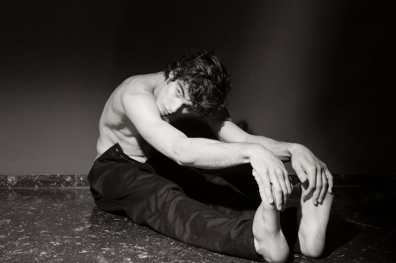 Monochrome portrait of a young adult male with dark hair, seated and shirtless, with arms stretched forward, against a dark background.