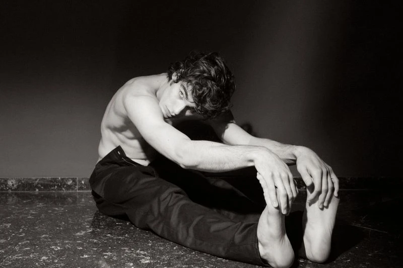 Monochrome portrait of a young adult male with dark hair, seated and shirtless, with arms stretched forward, against a dark background.