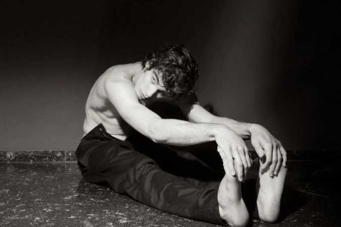 Monochrome portrait of a young adult male with dark hair, seated and shirtless, with arms stretched forward, against a dark background.