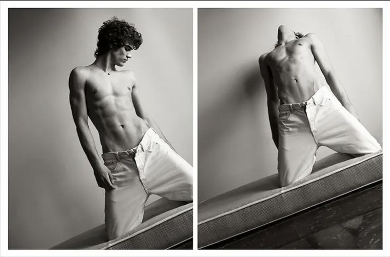Black and white portrait of a shirtless man with curly hair in light jeans, kneeling on a mattress against a plain background.
