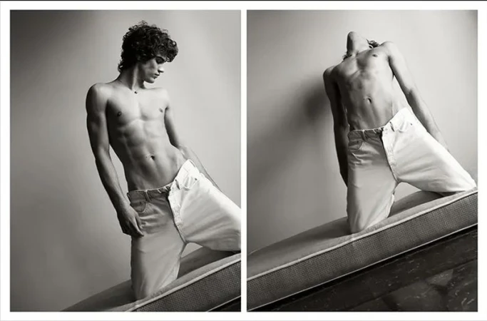 Black and white portrait of a shirtless man with curly hair in light jeans, kneeling on a mattress against a plain background.