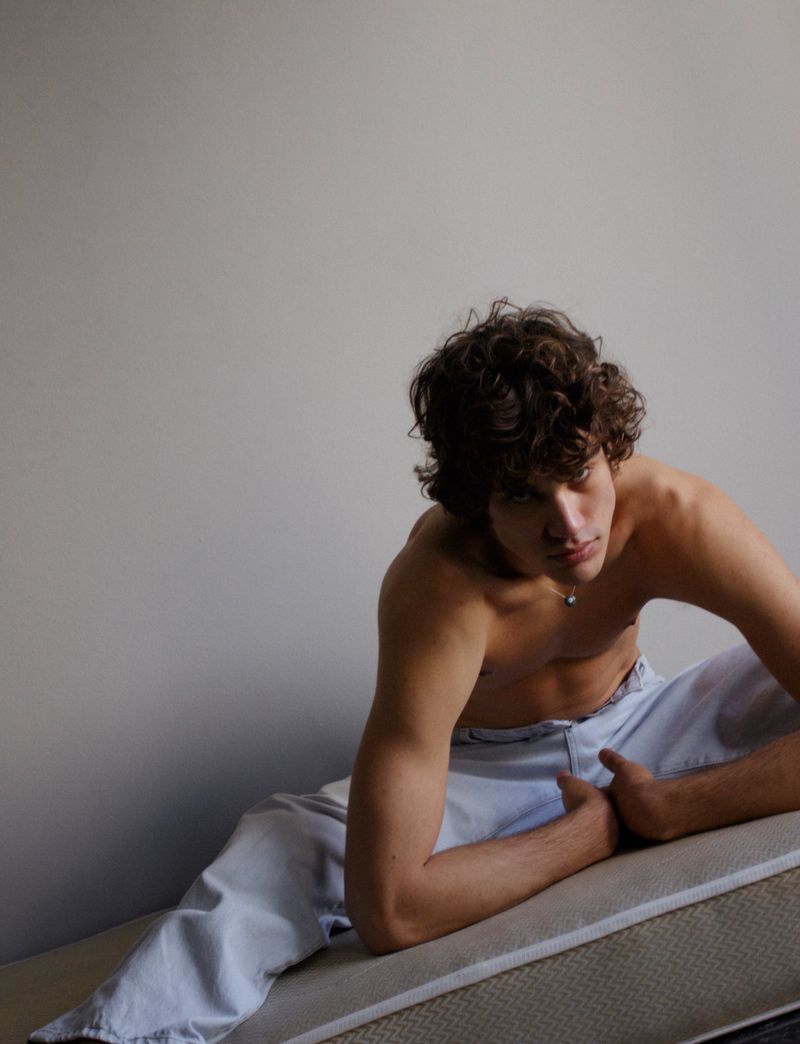 Man with curly hair posing shirtless in light-wash jeans, reclining indoors against a neutral wall, showing the fashion style in studio light.