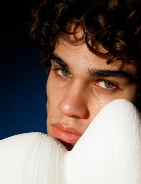 Close-up portrait of a young adult with curly hair, green eyes, and a white object, set against a dark blue background.
