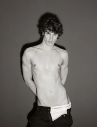 Shirtless young man with dark curly hair posing against a grey backdrop wearing unbuttoned dark trousers with white briefs visible.