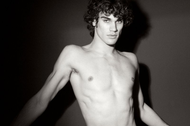 Monochrome portrait of a shirtless young man with dark curly hair posing against a dark background with strong shadow.