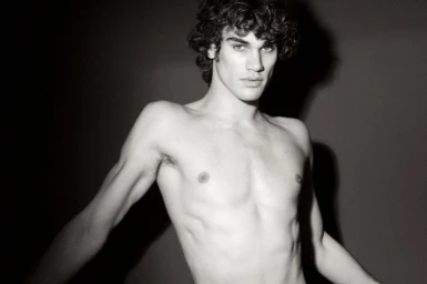 Monochrome portrait of a shirtless young man with dark curly hair posing against a dark background with strong shadow.