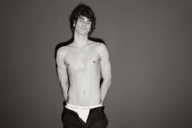 Shirtless young man with curly hair posing against a grey seamless studio background wearing low-slung trousers and branded briefs