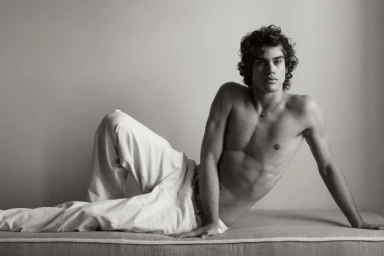 Shirtless young man with dark curly hair reclining on a mattress in white trousers against a plain studio wall.