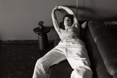 A shirtless man in relaxed white denim pants reclining on a dark sofa with arms raised overhead in a black-and-white editorial.