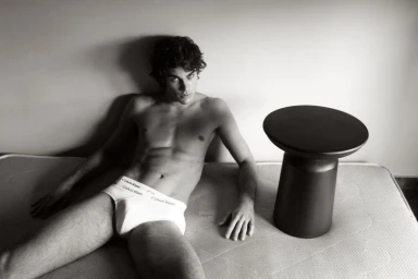 Shirtless man in white briefs reclining on a bare mattress next to a black stool against a plain wall.