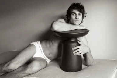 Man reclining in white branded briefs with his arms resting on a black sculptural side table.