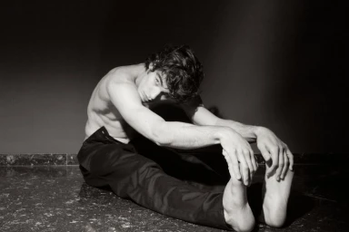 Shirtless young man stretching on a dark tiled floor while reaching for his bare feet in black and white.