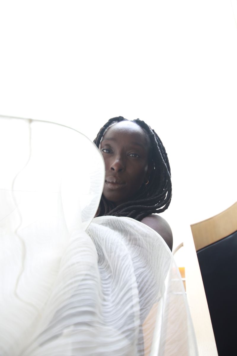 Portrait of an adult woman with dark hair and skin, partially veiled by sheer, horizontally striped white fabric against a bright background.
