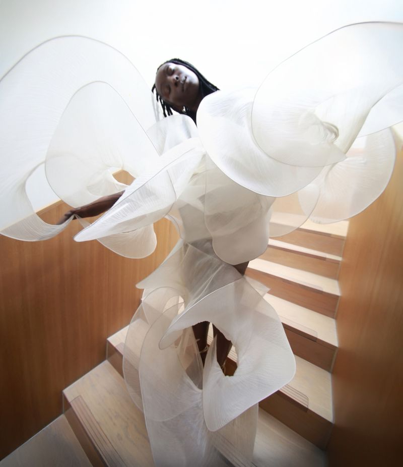 Eunice Olumide posing on a wooden staircase wearing an oversized, sculptural sheer white structured dress.