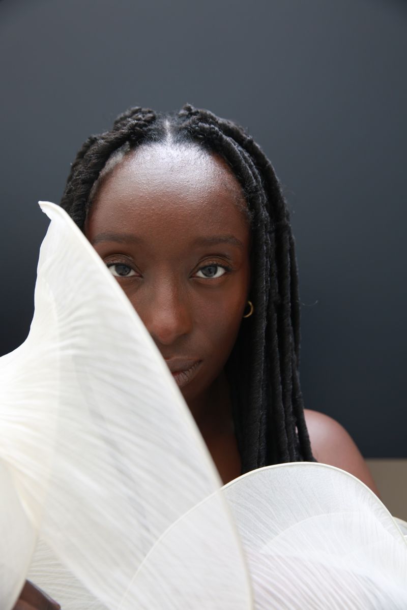Striking portrait of a Black woman with braided hair partially veiled by a flowing white fabric against a dark, neutral backdrop.
