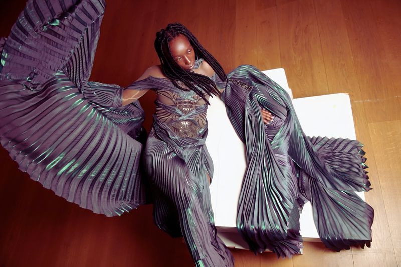 Fashion shot of adult female in a long, pleated teal-purple gown, lying on a white surface, with a wooden floor background.