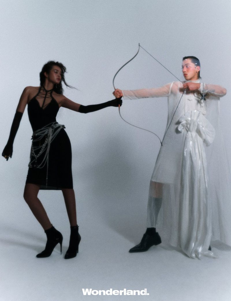 Two fashion models pose in a studio with a bow and arrow. One woman wears a black dress, and the other wears a sheer white dress.