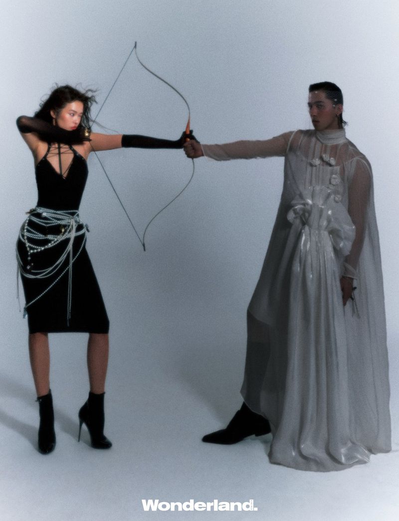 Fashion shot featuring a woman in a black dress holding a bow and arrow, facing a person in a white cape in a studio.
