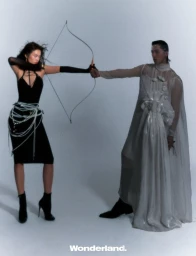 A woman in a black dress aims a bow and arrow at a man wearing a sheer floor-length gown against a pale studio backdrop.