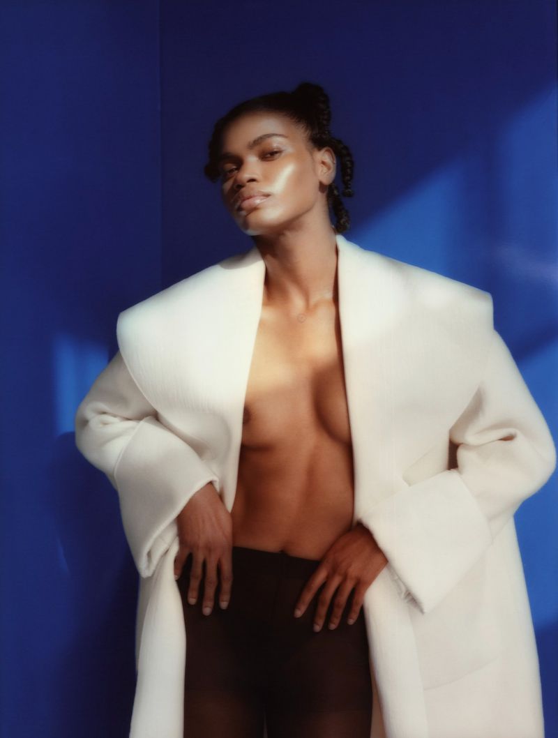 An adult female model with dark hair poses in a studio wearing a white coat and tights against a blue backdrop with shadow patterns.