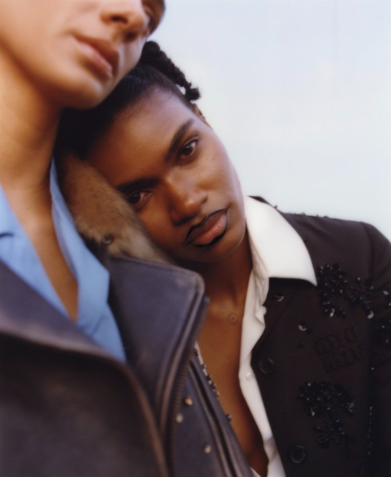 Close-up portrait of two adults, one resting head on the other's shoulder; one wears a black jacket, the other a blue shirt against a bright sky.