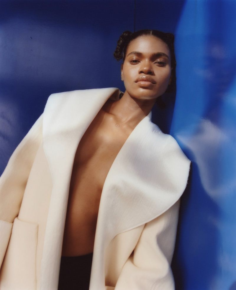 Fashion portrait of a young adult woman in an oversized cream coat, leaning against a blue wall with diffused lighting.