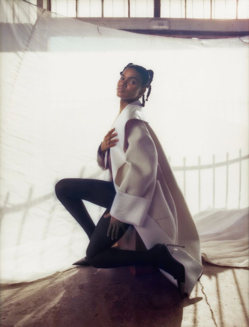 Elegant adult female model poses kneeling in a cream coat, black tights, and black high heels in a photo studio setting with soft lighting.