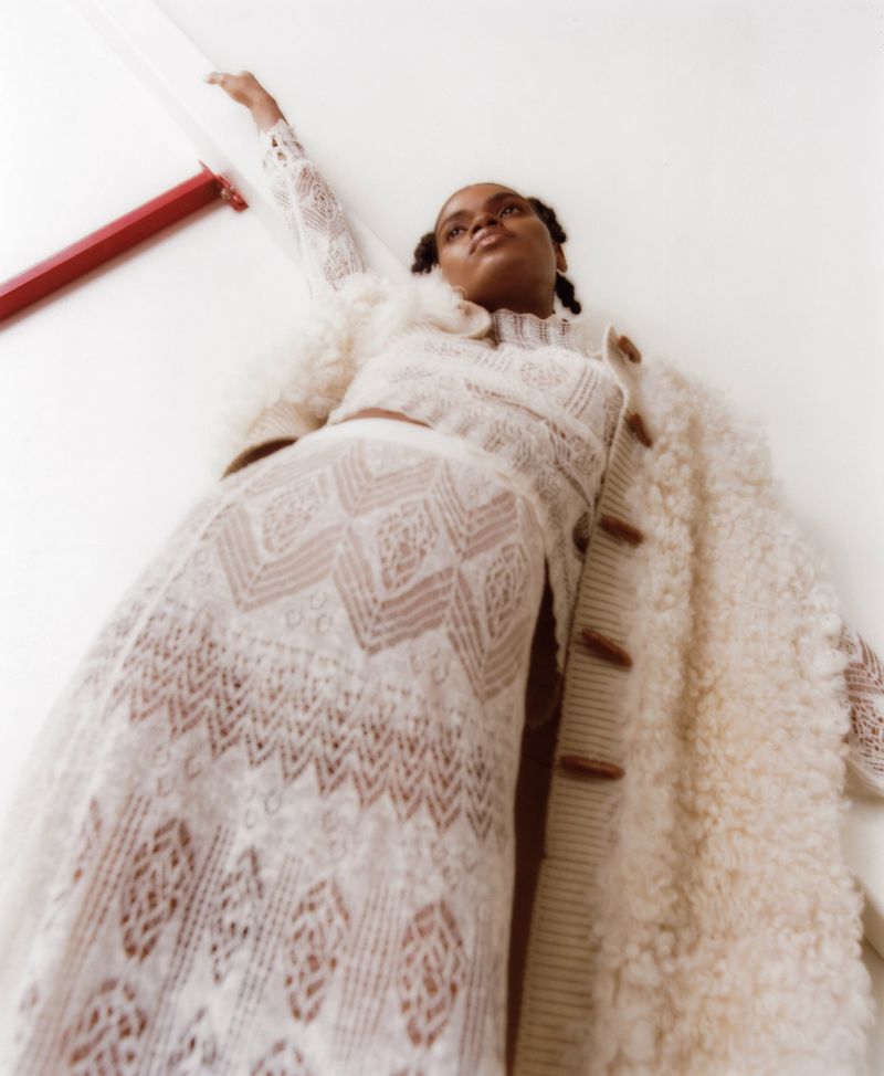 Portrait of an adult woman in a white, patterned outfit and a shaggy coat against a white background, captured from a low angle.