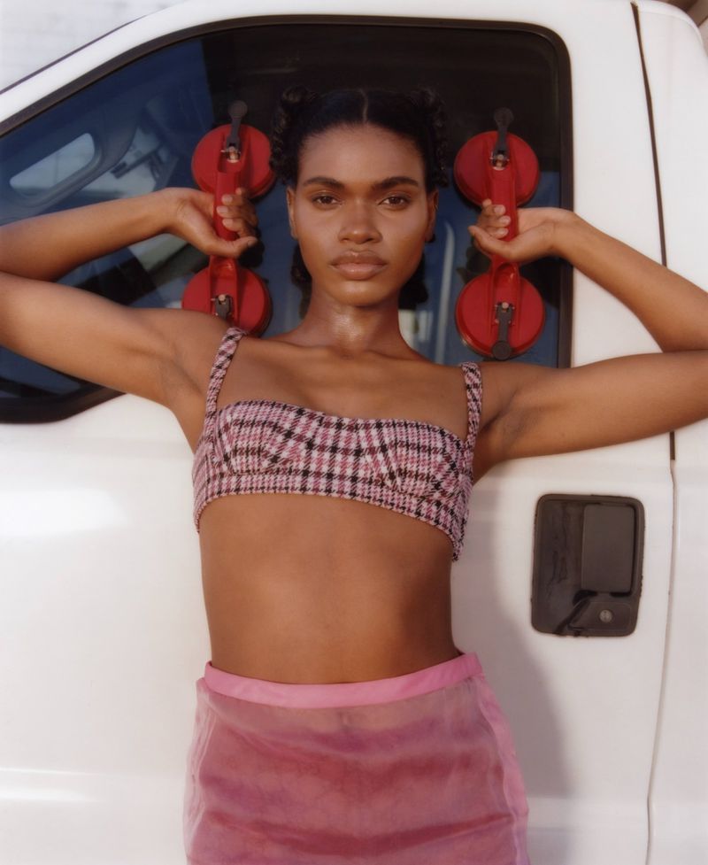 Portrait of a female model in a pink and black plaid top and pink skirt holding a red suction cup tool in front of a white truck.