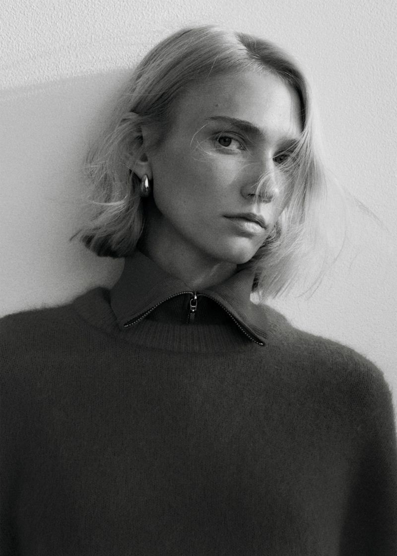 Monochrome portrait of a blonde woman with short hair wearing a zip-up sweater, posing indoors against a light-colored wall. Fashion shot.